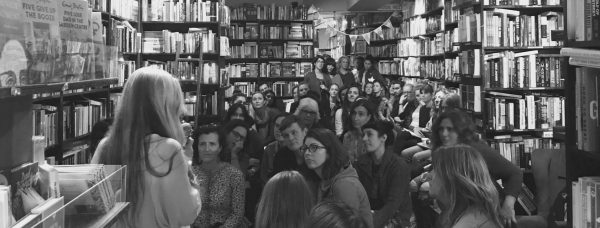 A black and white wholo of a bookstore filled with seated people watching a woman with long blonde hair holding a microphone who has her back to the camera.