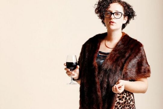 A person with dark curly hair standing against a plain light background, wearing leopard print and a brown fur sole, holding a glass of red wine.