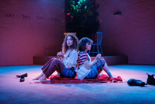 Two young women sit back to back on a theatre stage under blue and pink lights